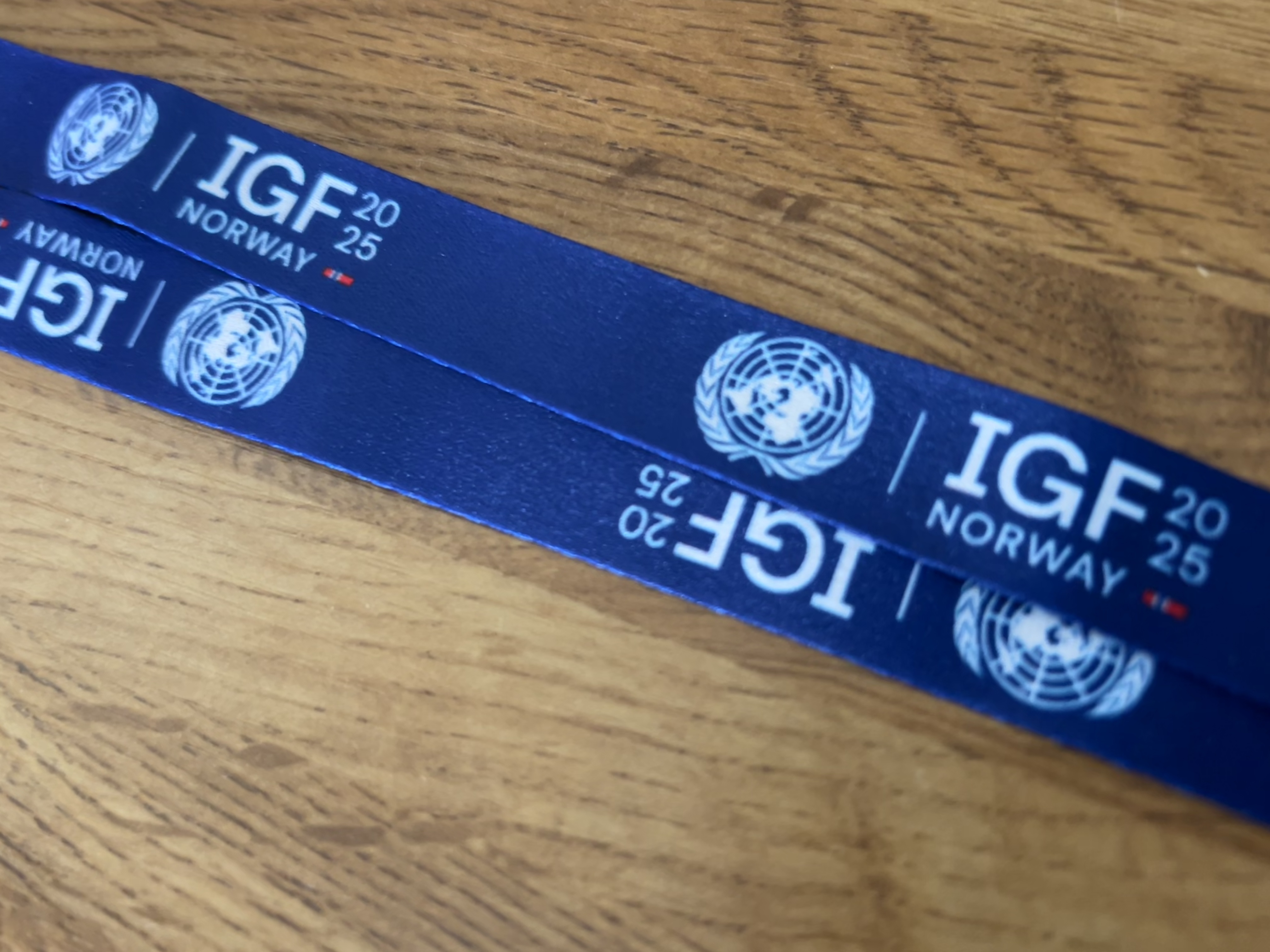 A blue lanyard from IGF 2025 in Norway, featuring the United Nations emblem and the text “IGF 2025 Norway” repeatedly printed along its length, lying on a wooden surface.