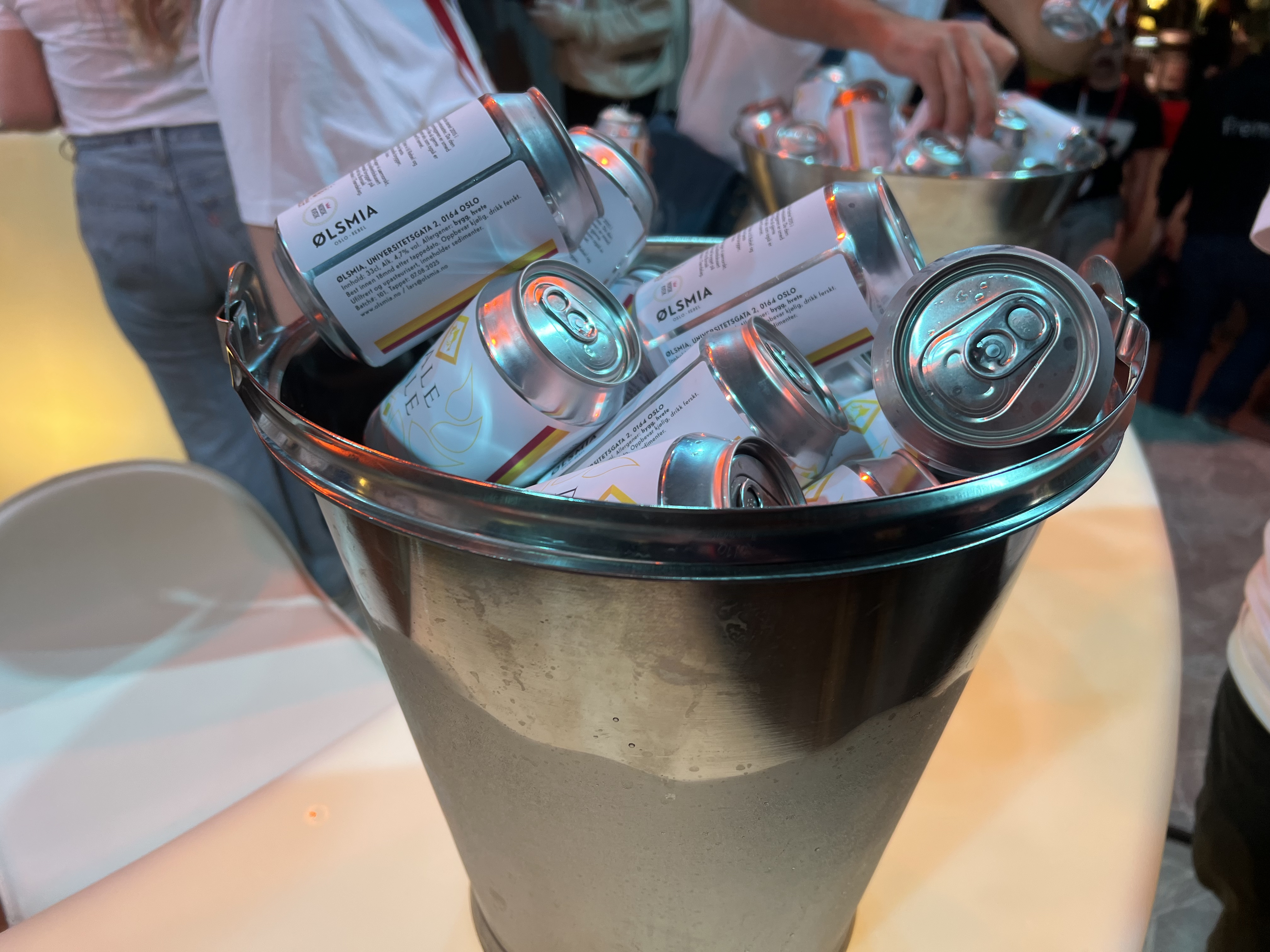 A metal bucket filled with several silver beer cans labeled “Ølsmia.” The bucket is placed on a table, and people are standing around in the background, some reaching for cans.
