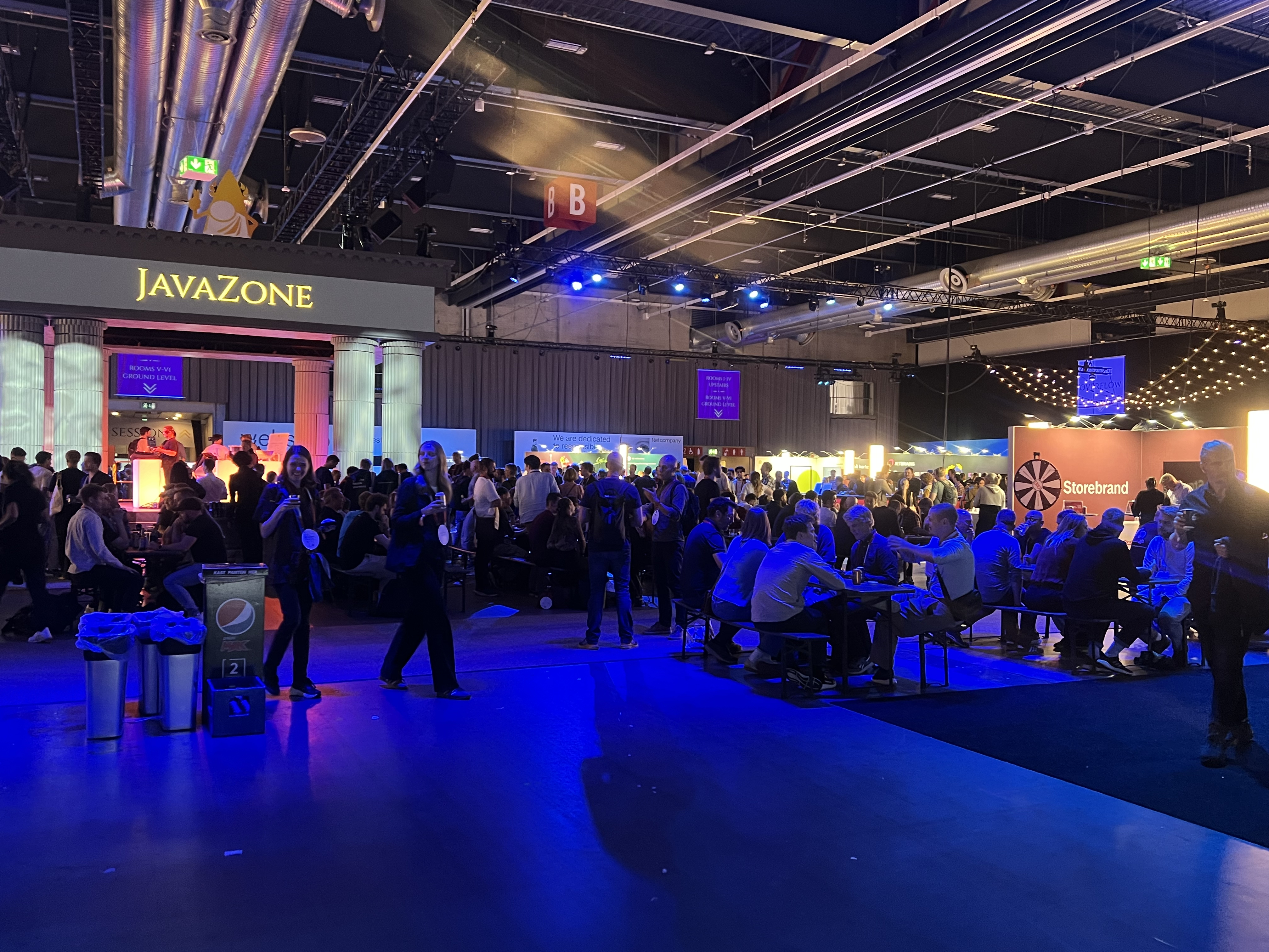 A large convention hall filled with people sitting at tables and walking around. On the left, there is a stage with a “JavaZone” sign above it, and on the right are various booths, including one from Storebrand. The lighting is dim with blue and purple spotlights, creating a lively atmosphere.