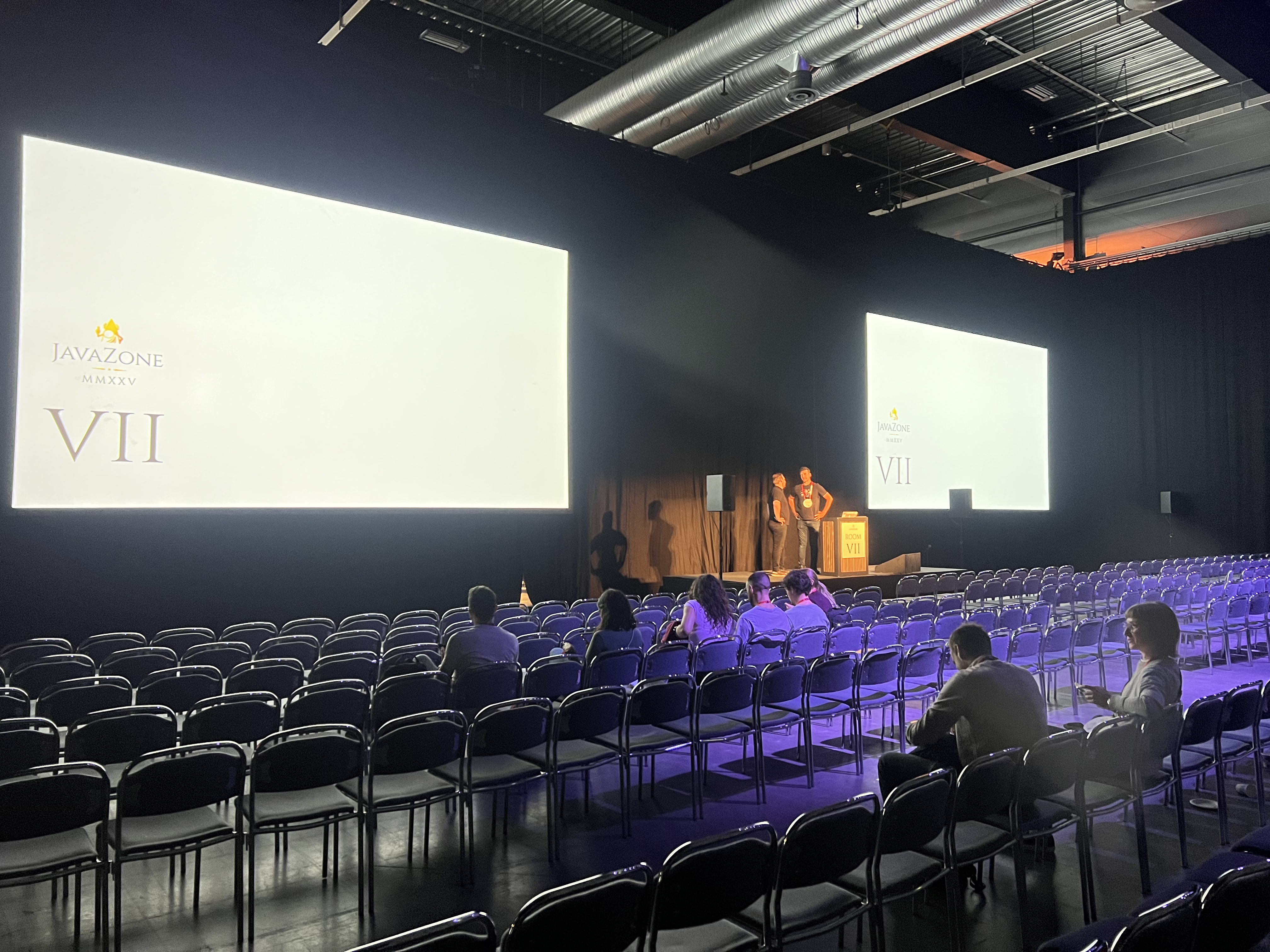 A large presentation room with rows of empty chairs facing two big screens displaying the JavaZone logo and the Roman numeral VII. Two speakers stand on stage at a podium preparing for a talk, while a few attendees sit scattered in the audience. The room is dimly lit, with focused stage lighting.