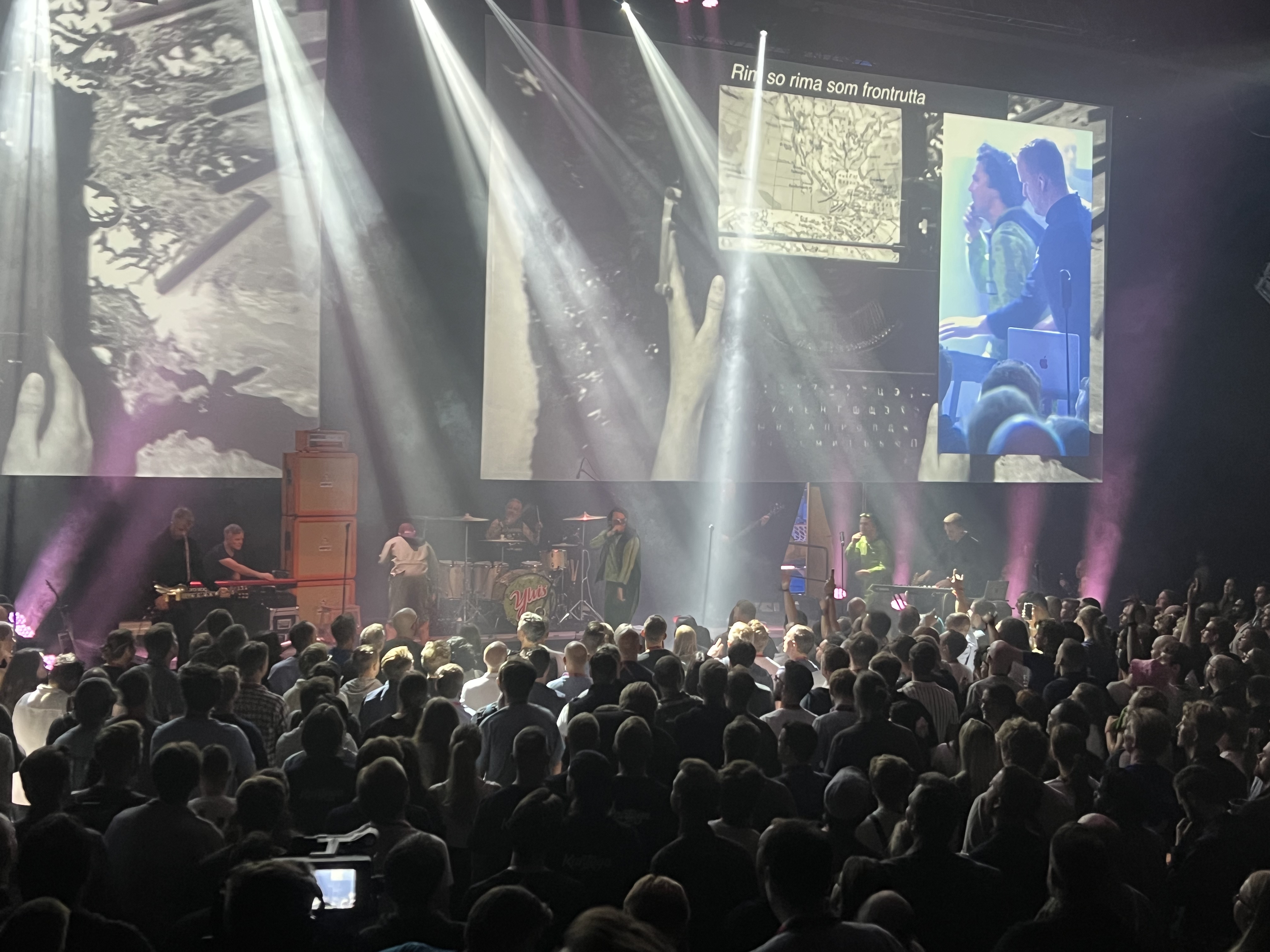 A concert stage with bright lights and large screens, where the Norwegian duo Ylvis is performing in front of a crowd. The audience is gathered in a dark hall, illuminated by colorful stage lighting.